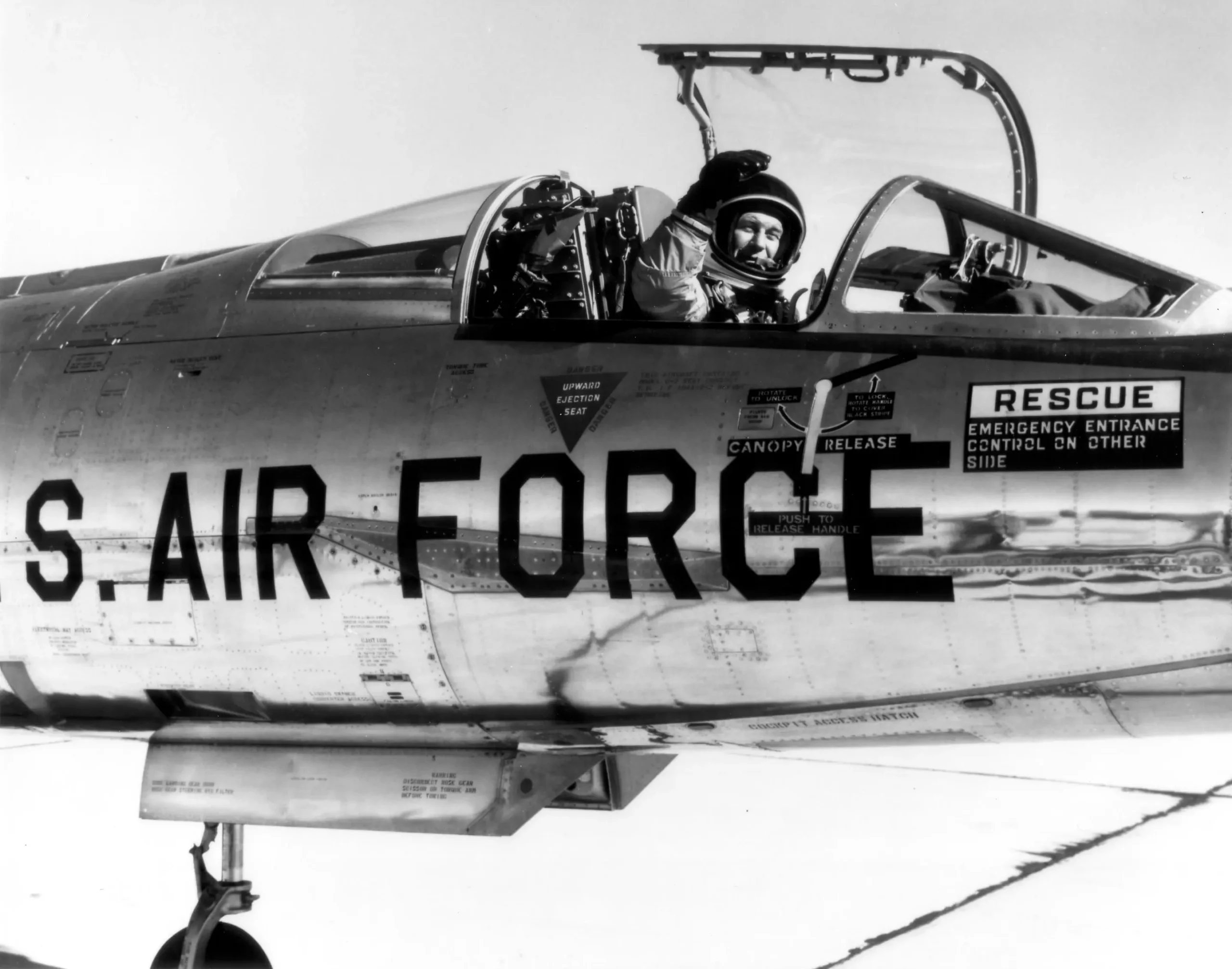 Black-and-white image of a pilot in a U.S. Air Force jet cockpit, wearing a flight suit and helmet. The open canopy and visible aircraft markings add detail.