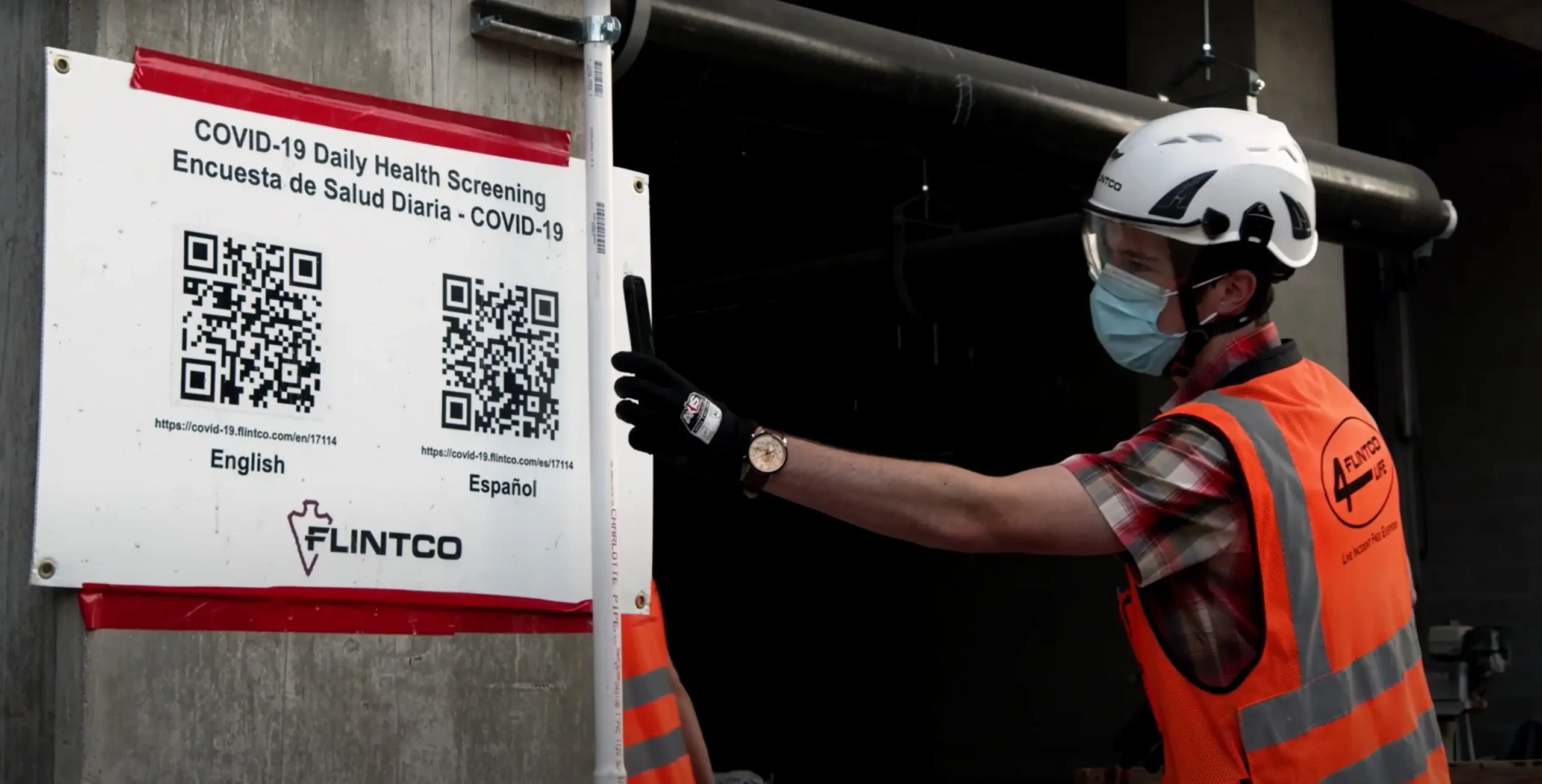 A construction worker in a safety vest and helmet adjusts a pole near a COVID-19 health screening sign with QR codes. The scene conveys caution.