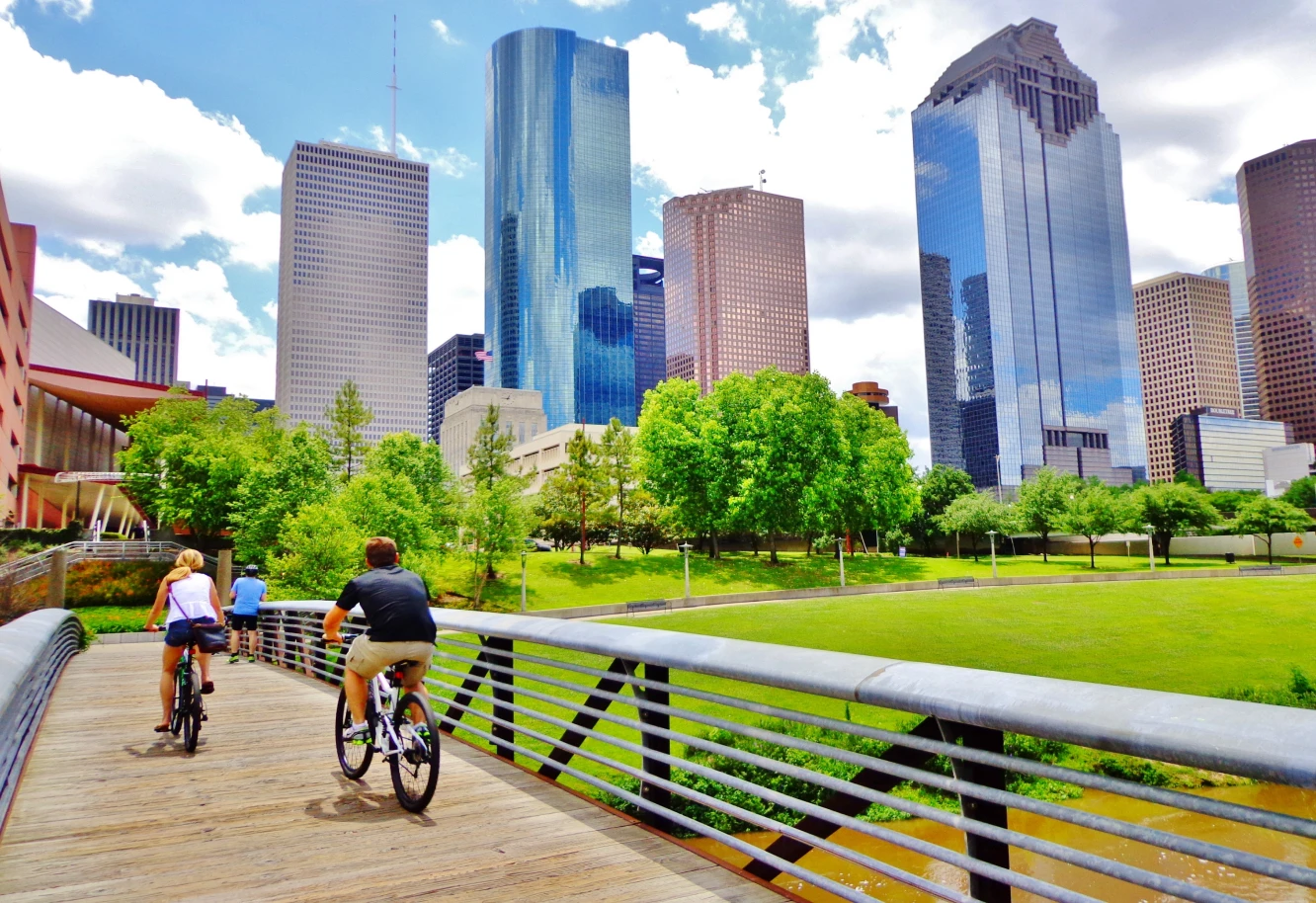 Three people ride bicycles on a wooden path surrounded by green trees, with a backdrop of modern skyscrapers under a bright blue sky in the city.