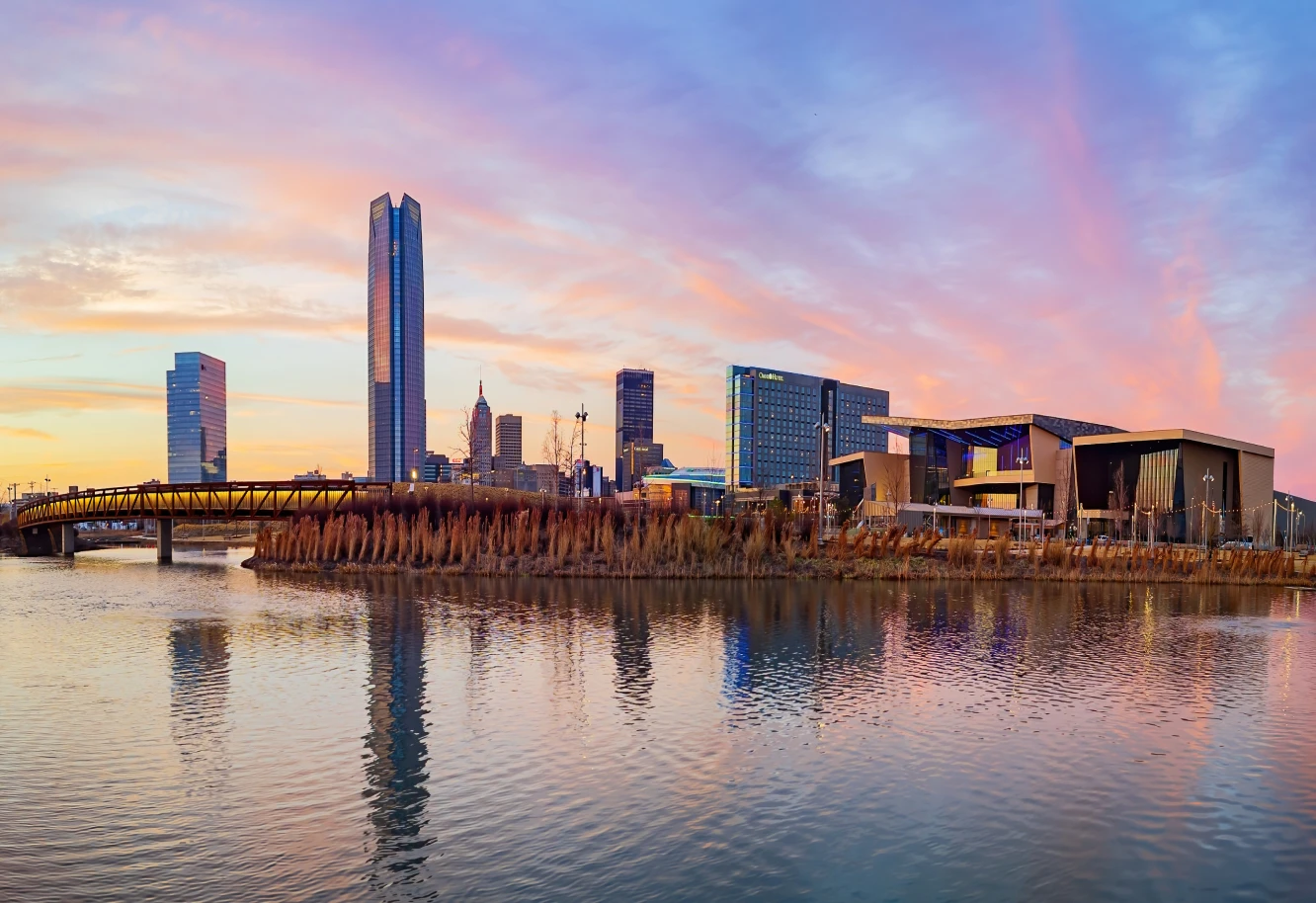 A city skyline at sunset with tall buildings and a modern bridge reflected in a calm river. The pink and orange sky creates a serene atmosphere.