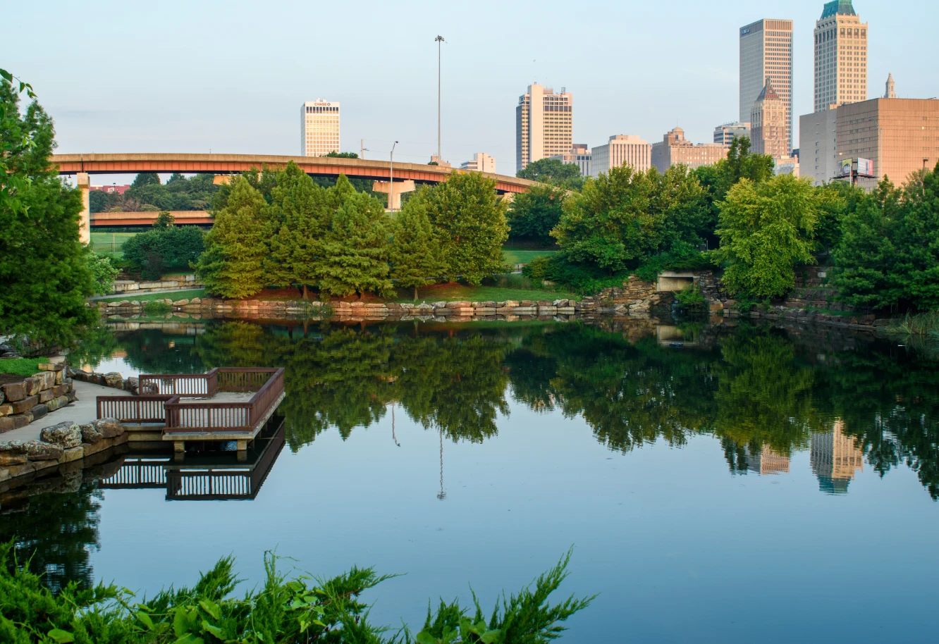 A serene lake reflects lush green trees and an urban skyline with tall buildings under a clear sky, conveying a calm, peaceful urban oasis.
