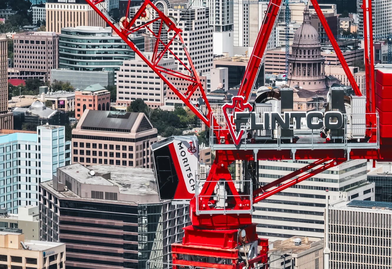 Red construction crane with "FLINTCO" sign looms over a cityscape of high-rise buildings and the dome of a historic capitol building, conveying urban development.