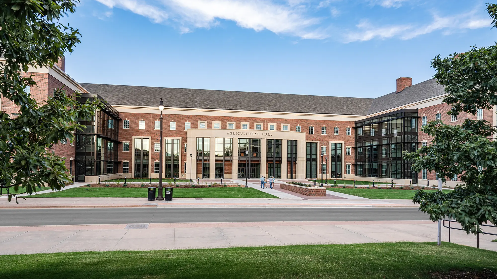 Brick building labeled "Agricultural Hall" with tall glass windows, surrounded by green lawns and trees. A clear, blue sky suggests a bright day.