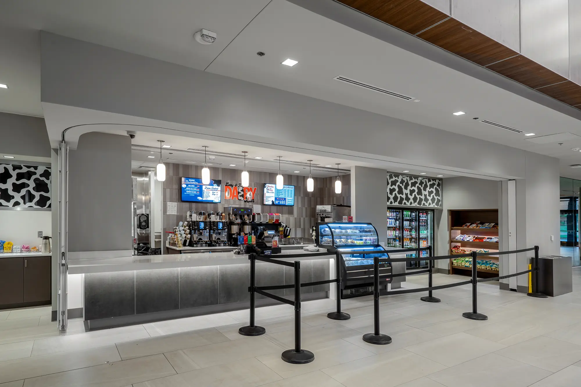 Modern snack bar with a coffee station, light fixtures, menu displays, and stocked shelves of beverages and snacks. Clean, minimalist design.