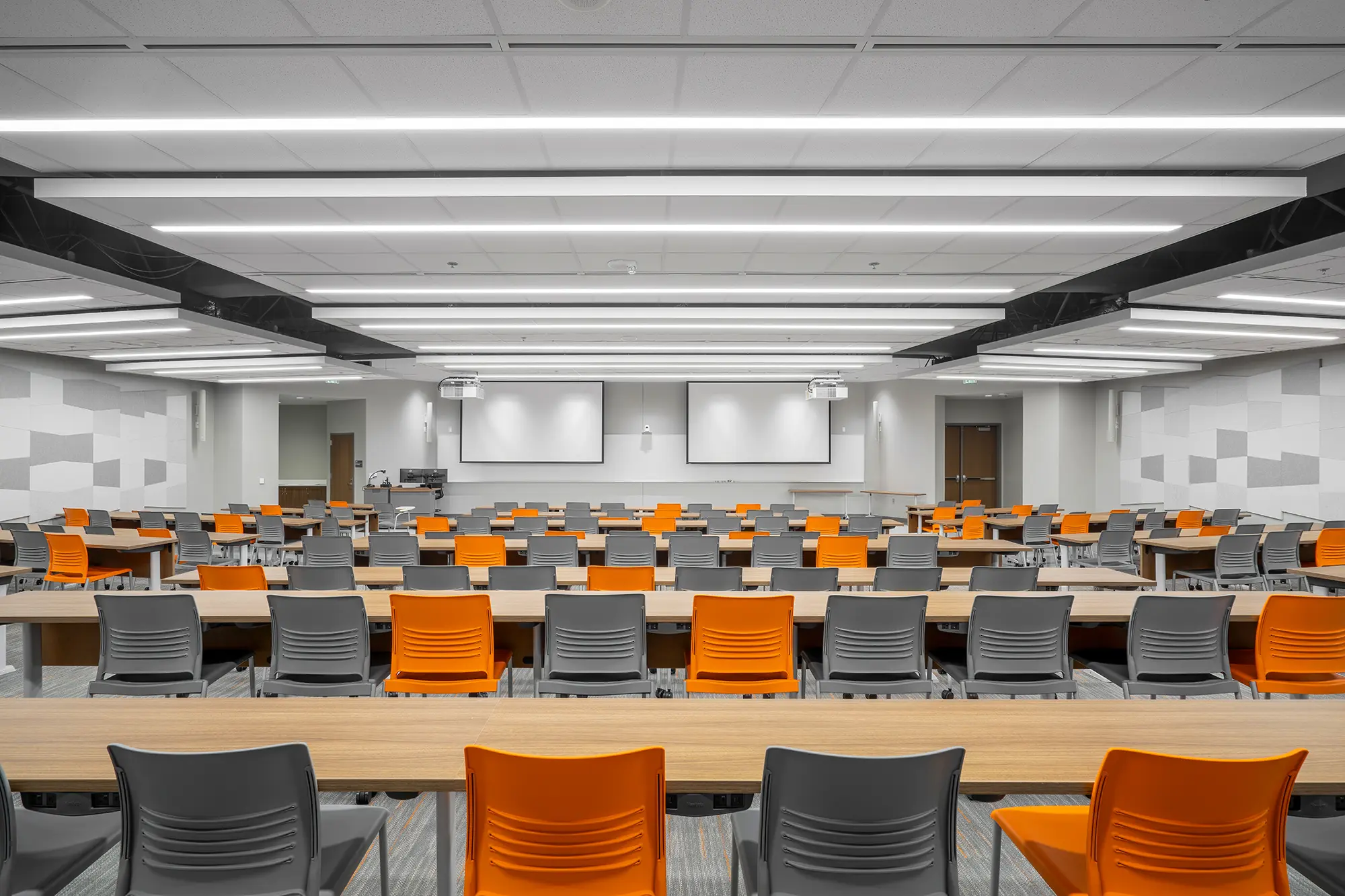 Modern lecture hall with rows of gray and orange chairs, wooden desks, and bright lighting. Three screens at front convey a professional tone.