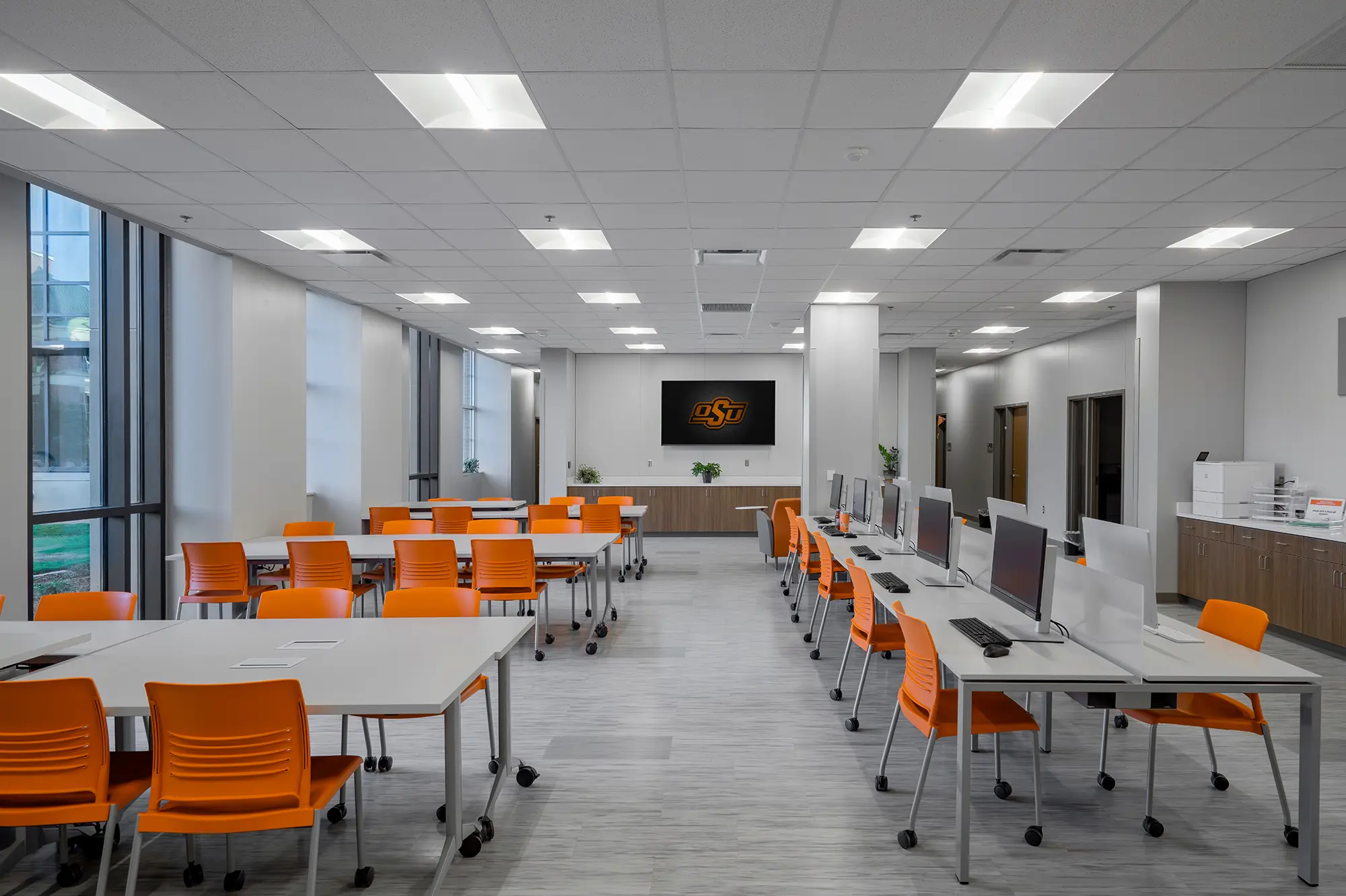 Modern computer lab with orange chairs, white desks, and several computers. Bright, well-lit space with a screen displaying a logo on the wall.