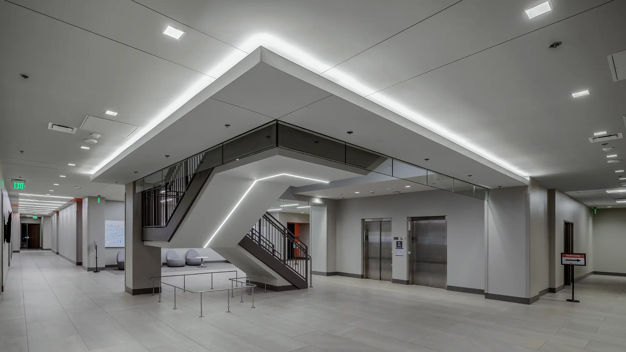 Spacious modern lobby with bright white lighting, sleek staircase, and two elevators. Minimalist design conveys a clean, professional atmosphere.