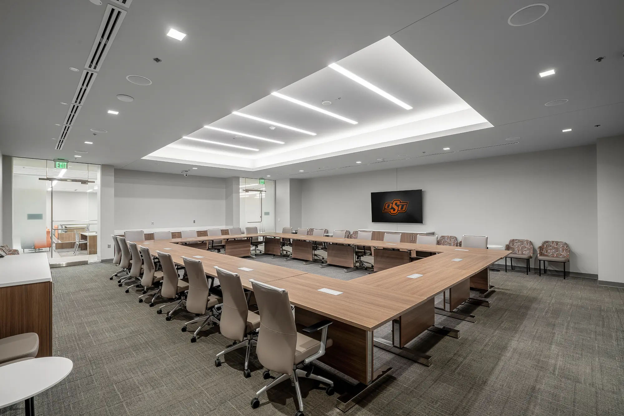 Modern conference room with U-shaped wooden table, beige swivel chairs, and a wall-mounted screen displaying a logo. Bright, minimalistic, professional ambiance.