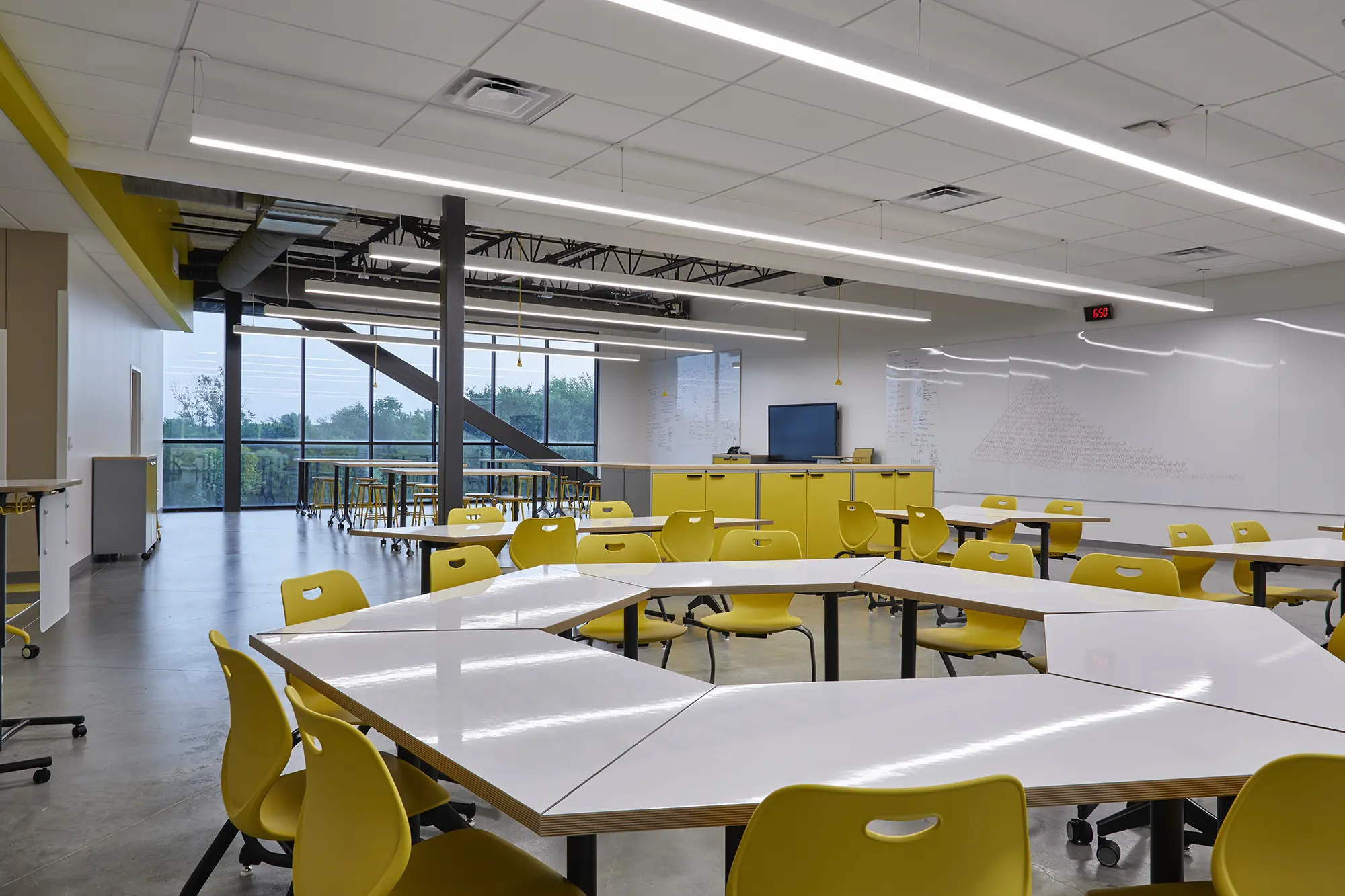 Spacious, modern classroom with yellow chairs, white hexagonal tables, large windows showing greenery, and bright overhead lighting, creating an inviting study environment.