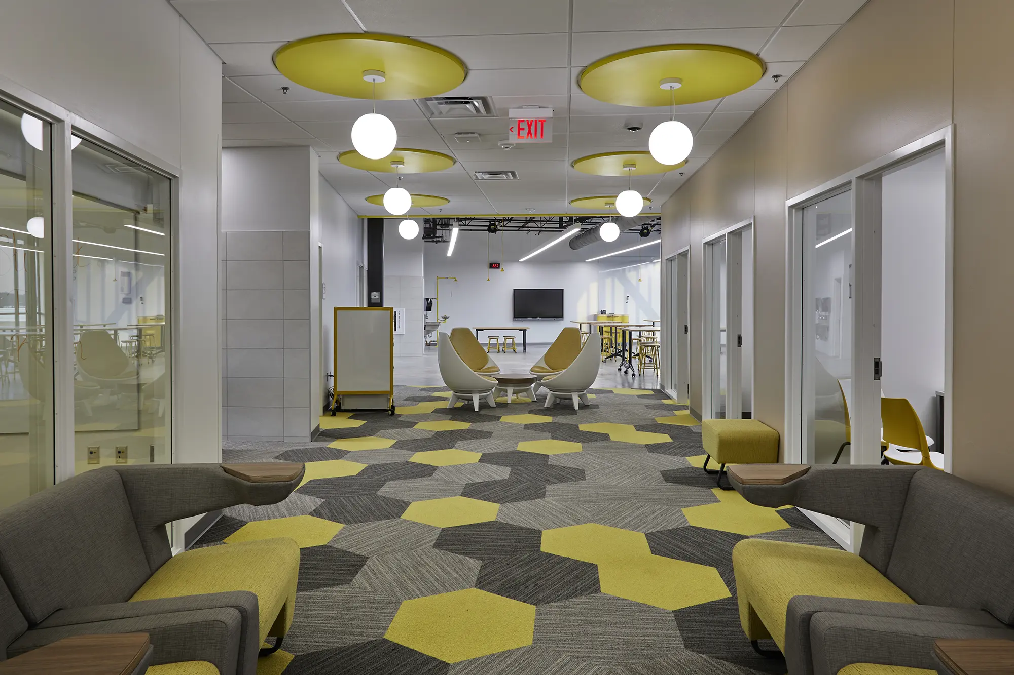Modern office lounge with hexagonal yellow and gray carpet, yellow seating, round white chairs, and circular ceiling lights, creating a vibrant, inviting atmosphere.