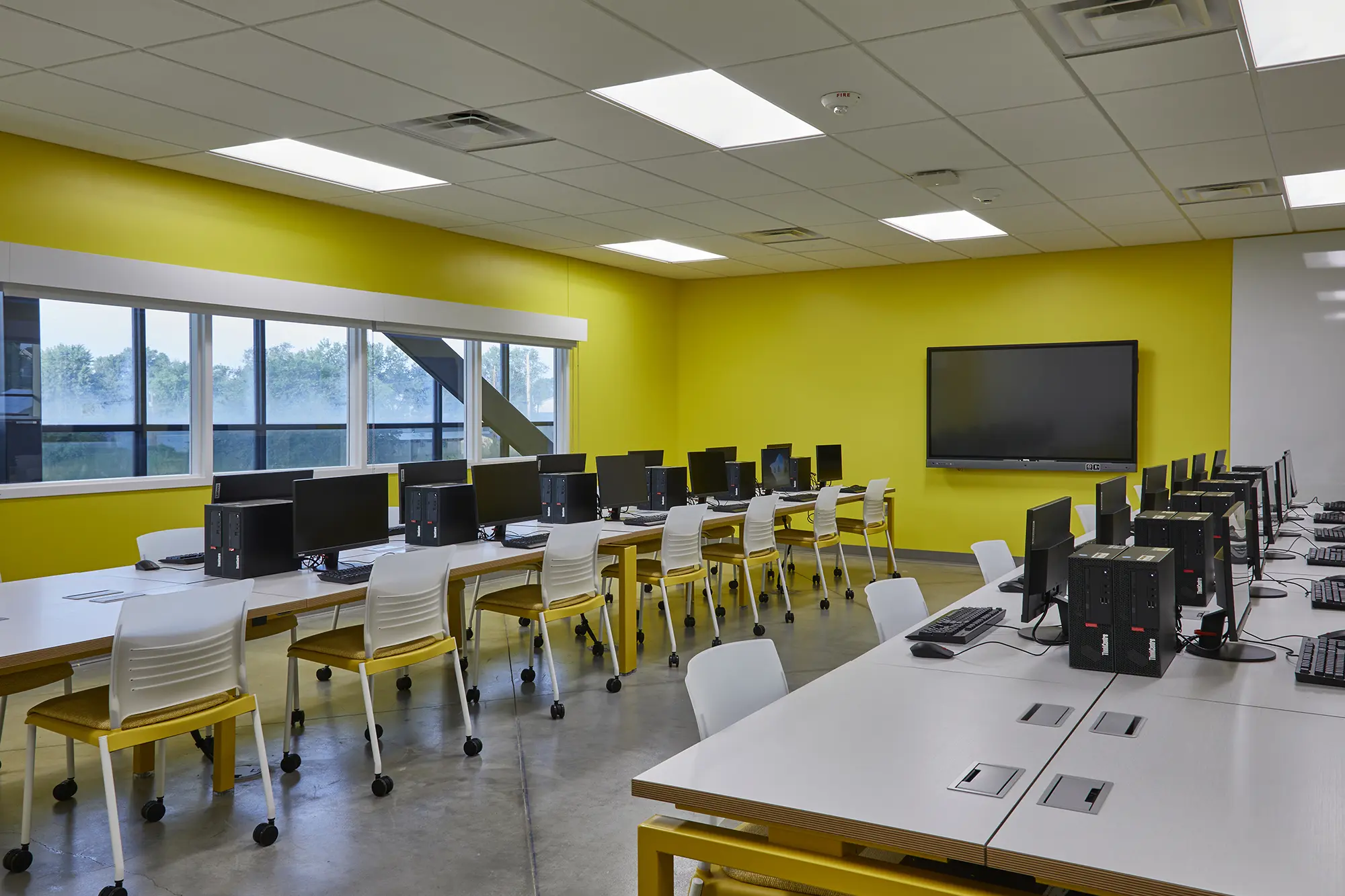 Modern computer lab with bright yellow walls, large windows, and rows of computers on white desks. The atmosphere is clean and tech-focused.
