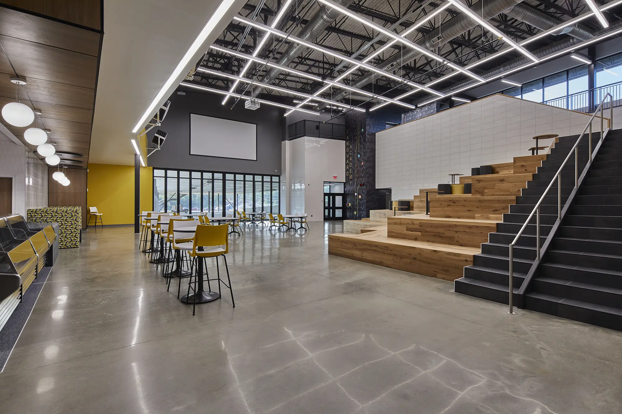 Spacious modern interior with high ceilings and geometric lighting. Features wooden bleacher seating, yellow chairs at tables, and large windows.