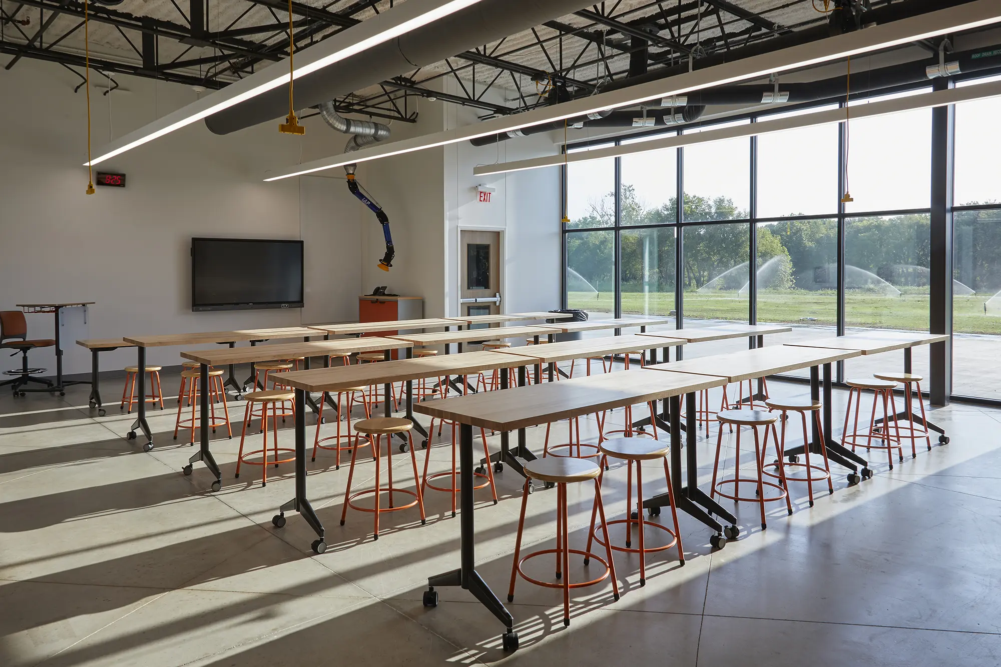 Modern classroom with long tables and orange stools under bright lights. Large windows reveal a green landscape, creating an open and inviting atmosphere.