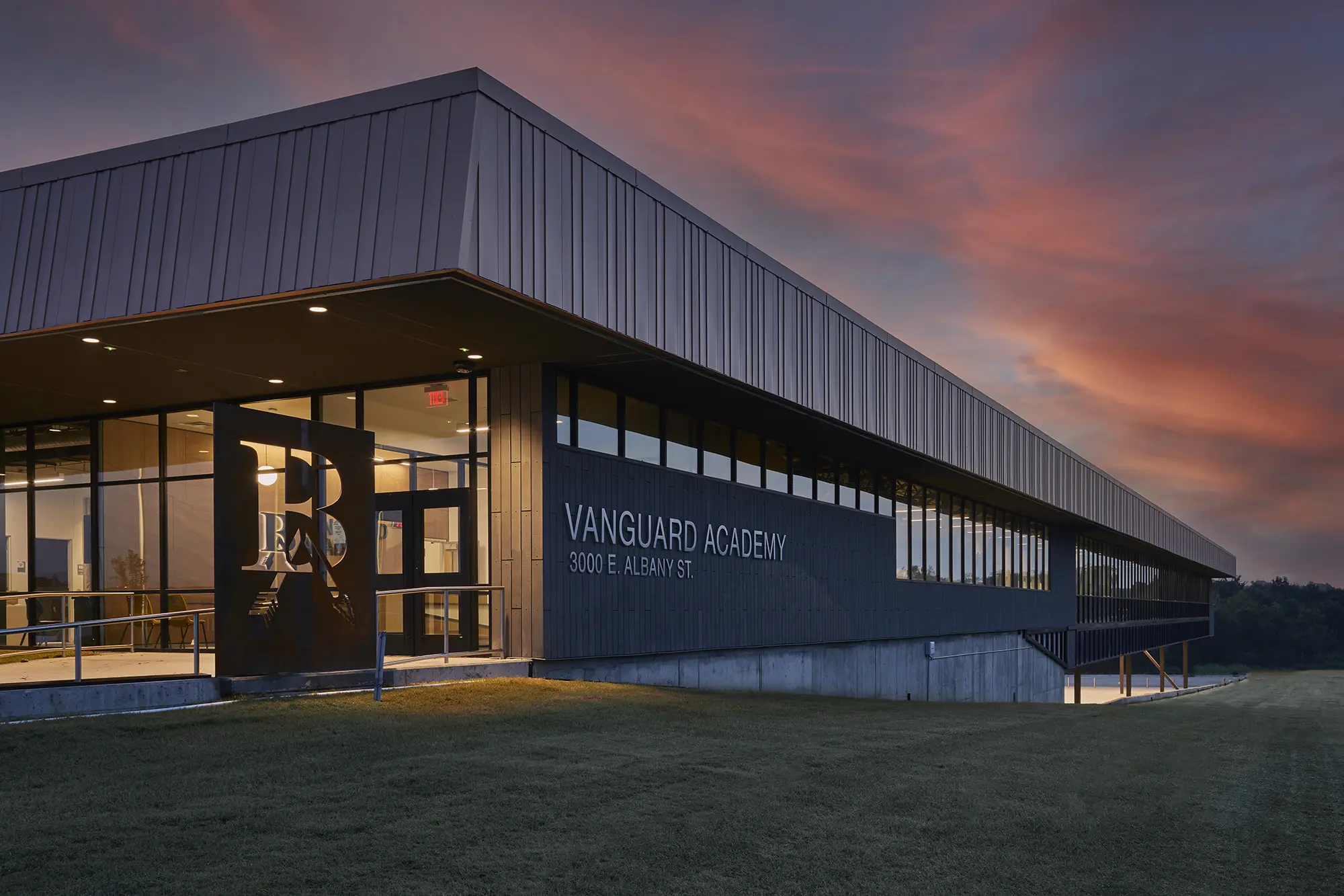 Modern building labeled "Vanguard Academy" at sunset, with a sleek design, large windows, and warm lighting, under a vibrant, colorful sky.