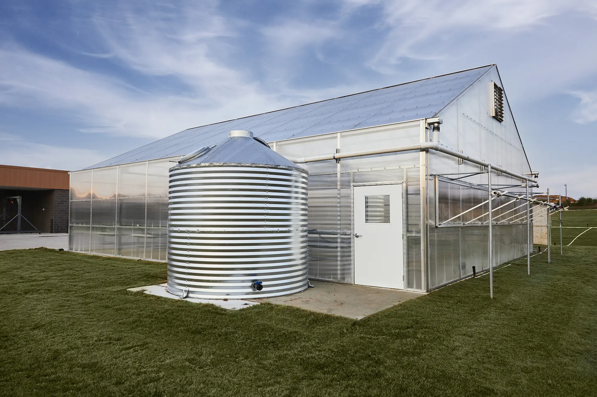 A modern greenhouse with a large metallic water tank stands on a grassy lawn under a partly cloudy sky, conveying a sense of innovation and sustainability.