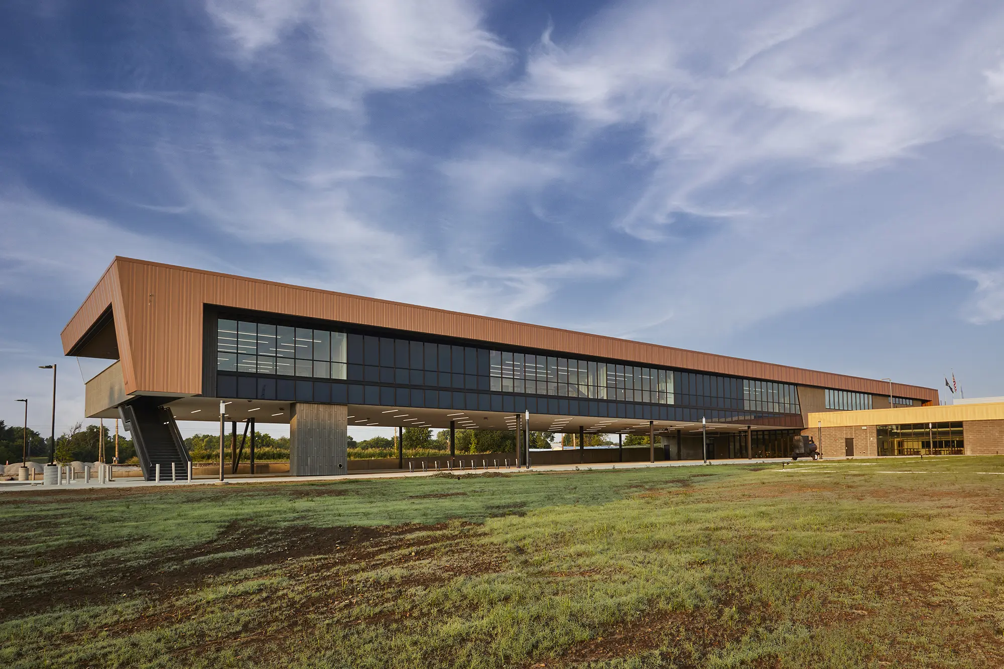 Modern, angular building with a copper roof and large glass windows, elevated on pillars. Set against a clear blue sky with wispy clouds, surrounded by grass.