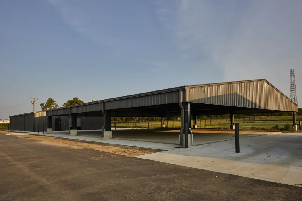 Large, open-walled metal pavilion with a gabled roof and concrete foundation, set in a grassy area under a clear blue sky, conveying spaciousness.