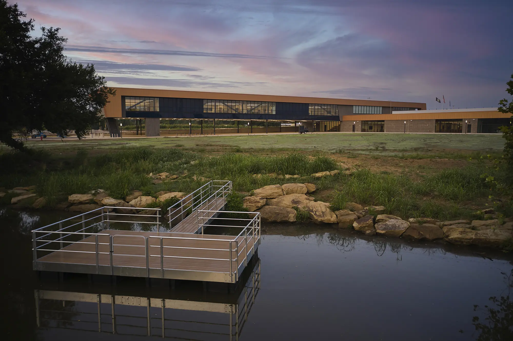 Modern building with large windows at sunset, reflected in a calm pond with a floating deck in the foreground, surrounded by rocks and greenery.