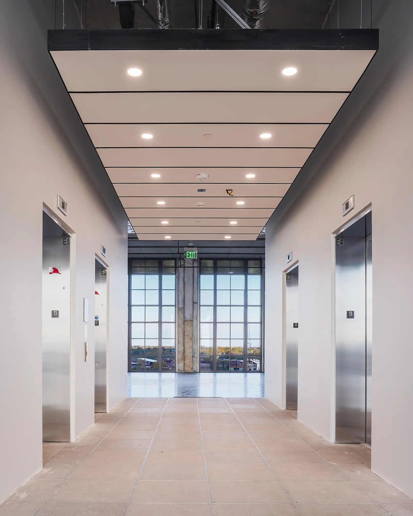 Hallway with four modern elevators, two on each side, under a sleek ceiling with recessed lights. Large window at the end reveals a distant landscape.