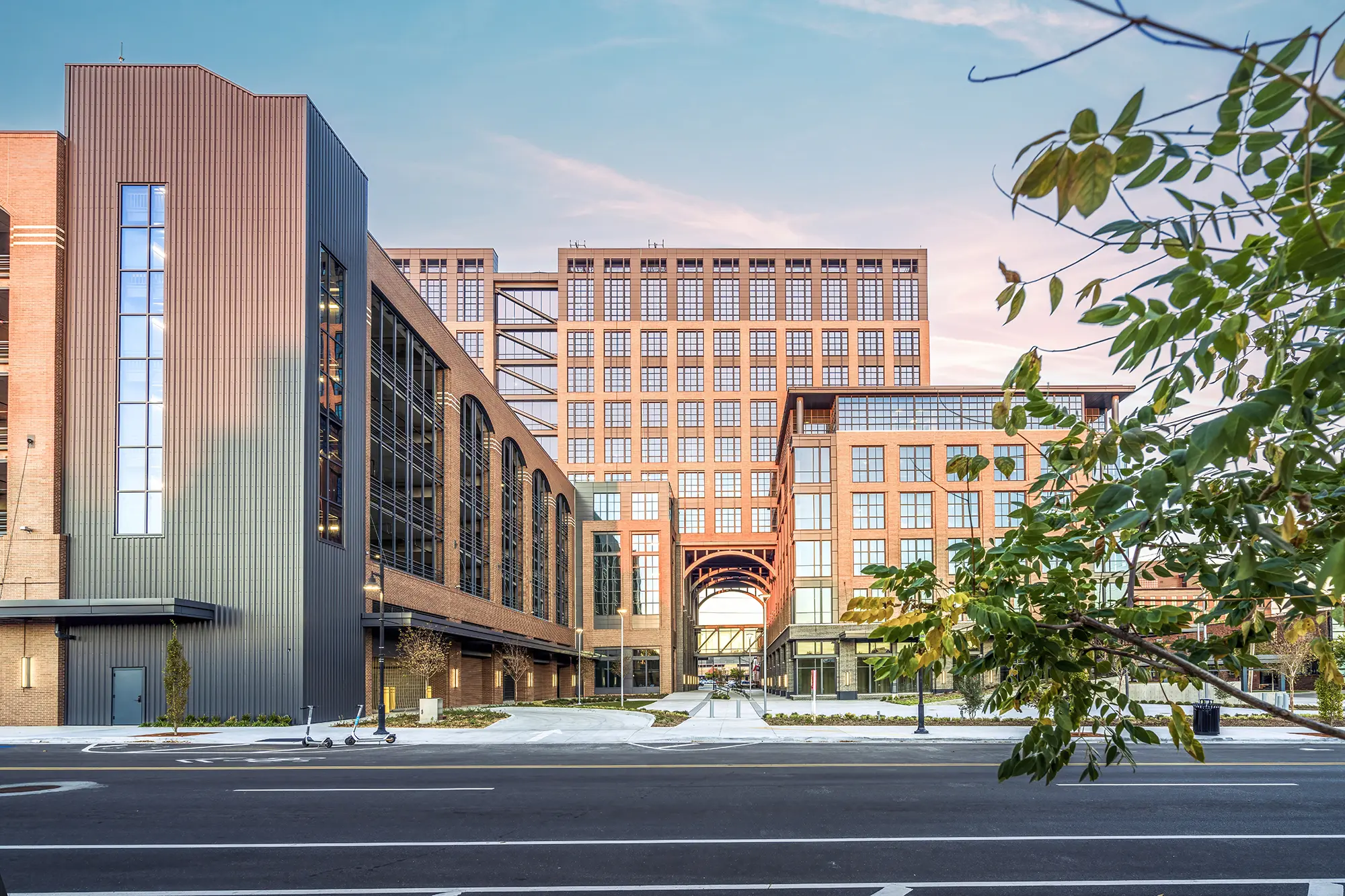 Modern urban building complex with brick and metal facades under a vibrant sunset sky. Leafy foreground and empty street add tranquility.