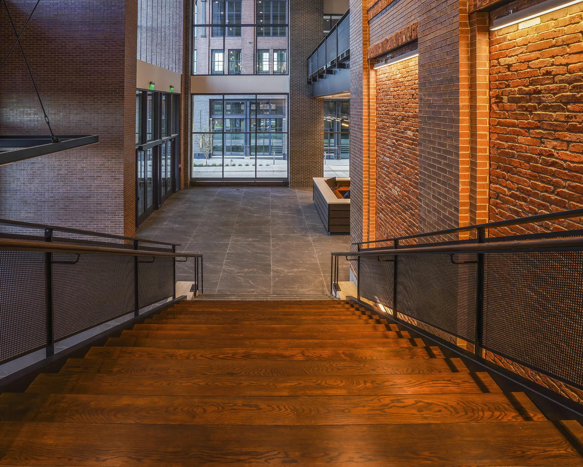 Spacious industrial lobby with warm lighting, featuring wooden stairs, exposed brick walls, large windows, and a stone floor, creating an inviting atmosphere.