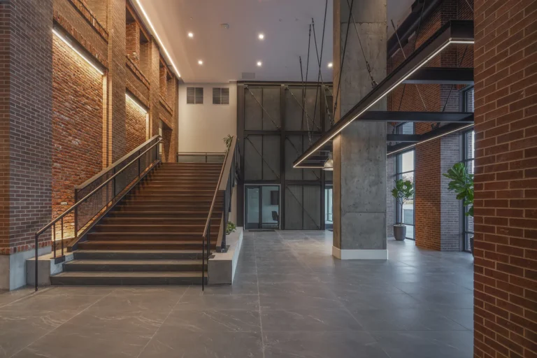 Spacious modern lobby with exposed brick walls, large staircase, and industrial lighting. Potted plants add greenery, creating a warm, inviting atmosphere.