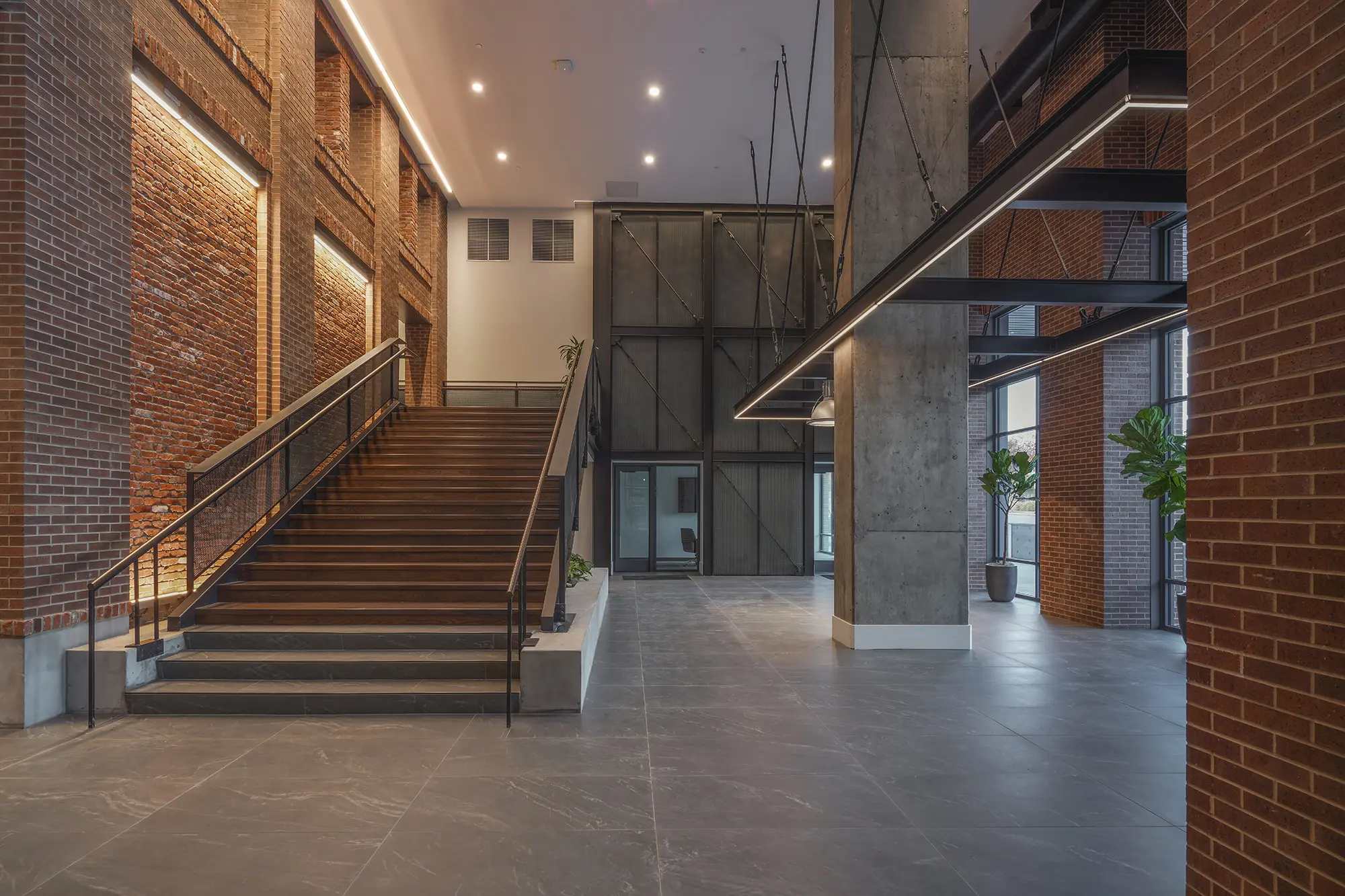 Spacious modern lobby with exposed brick walls, large staircase, and industrial lighting. Potted plants add greenery, creating a warm, inviting atmosphere.