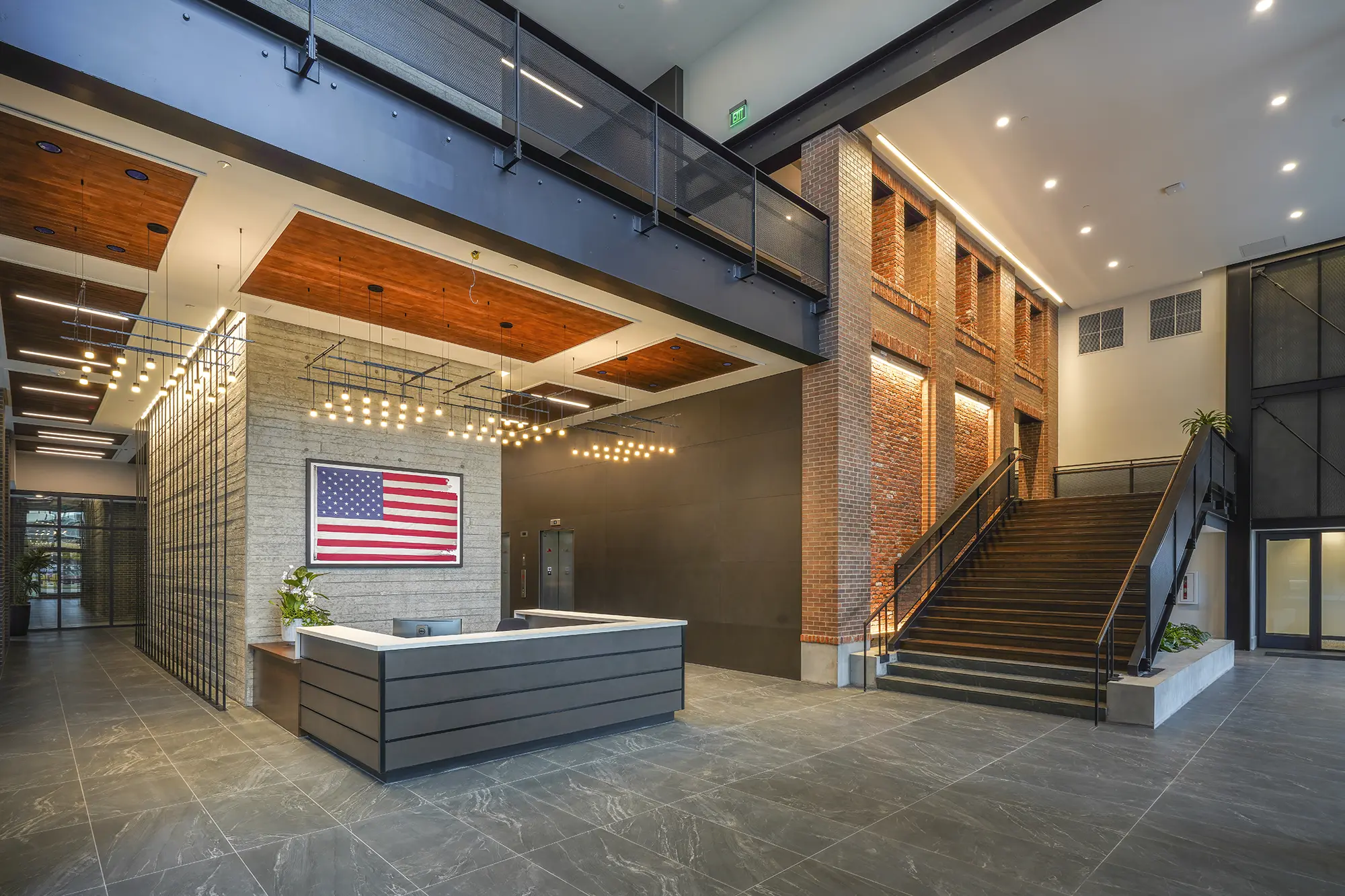 Modern lobby with brick walls, a sleek reception desk, and an American flag. Spacious, with warm lighting, a staircase, and industrial accents.