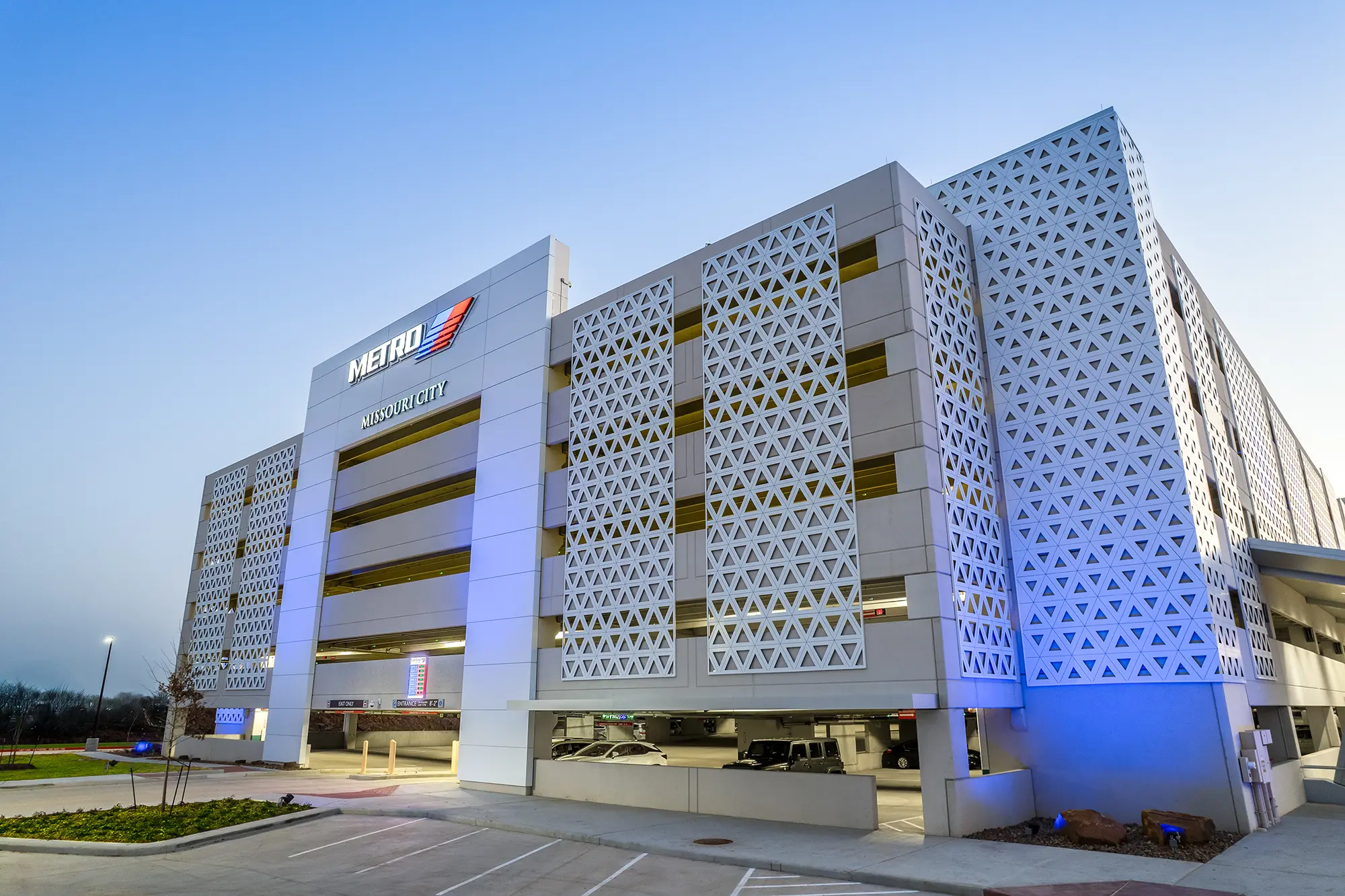 Modern parking garage at dusk, featuring geometric lattice panels and "Metro Missouri City" signage. Soft blue lighting creates a sleek, urban ambiance.