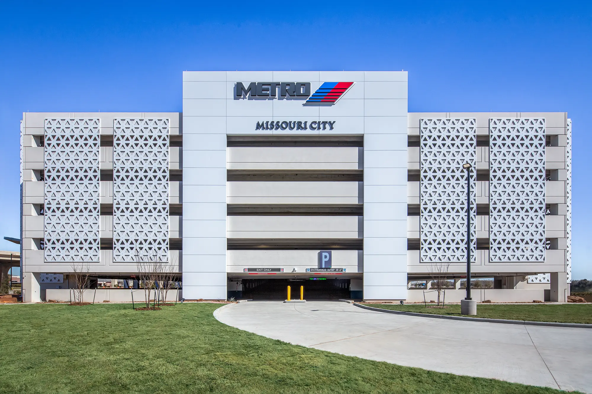 Modern parking garage with bold “Metro” and “Missouri City” signage. Features geometric facade panels, a central entrance, and a surrounding grassy area.