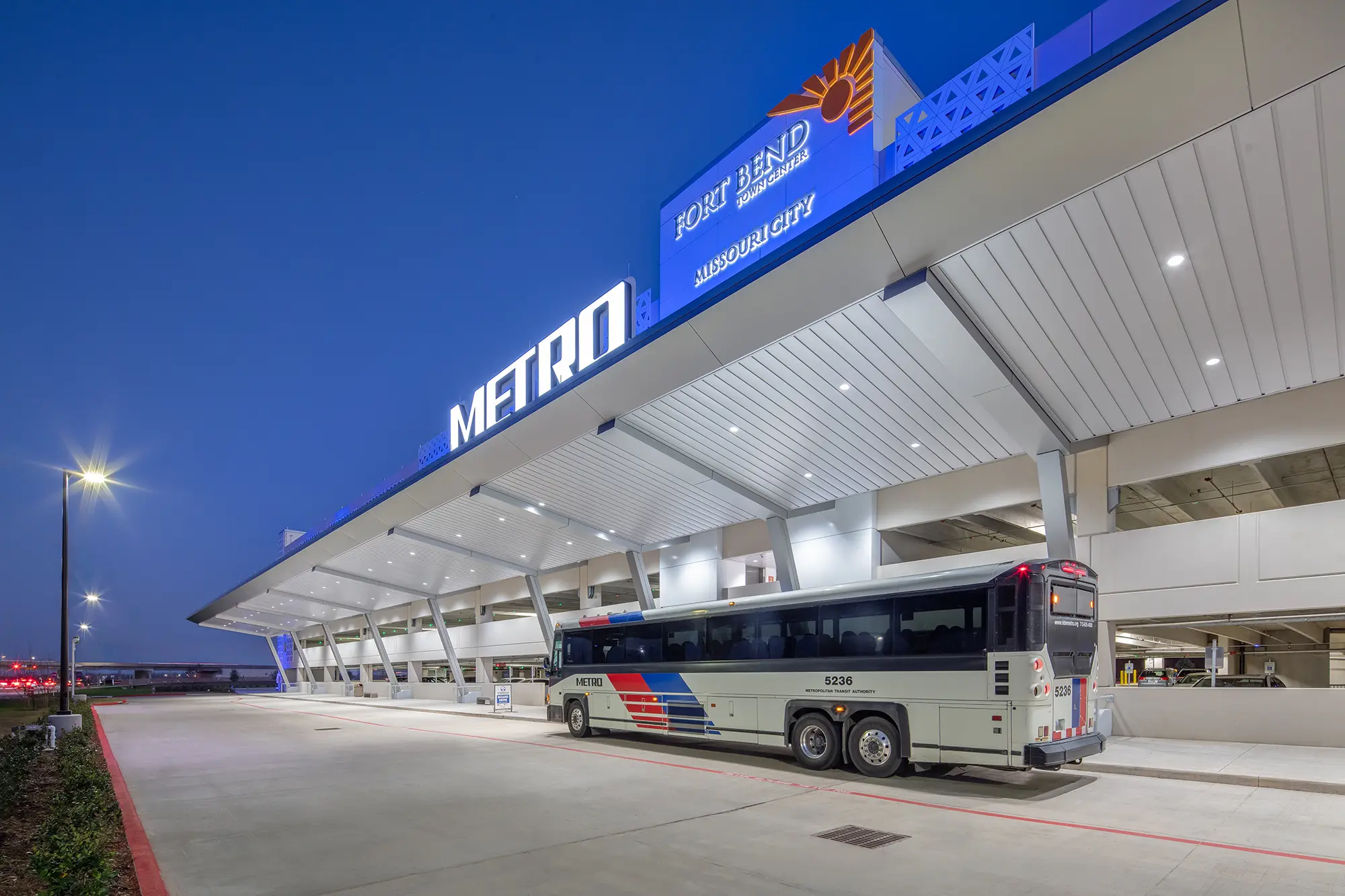 A METRO bus is parked at a modern transit center beneath a well-lit canopy at dusk. Signage reads "Fort Bend" and "Missouri City." The scene is calm and orderly.