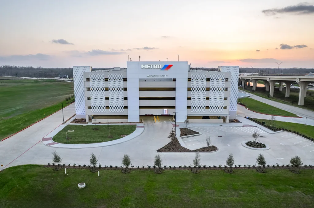 Aerial view of a modern, multi-level parking garage with METRO signage at sunset. The foreground features a neat driveway and landscaped greenery.