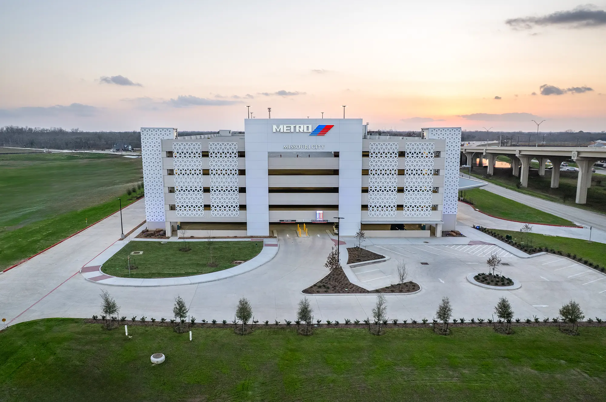 Aerial view of a modern, multi-level parking garage with METRO signage at sunset. The foreground features a neat driveway and landscaped greenery.