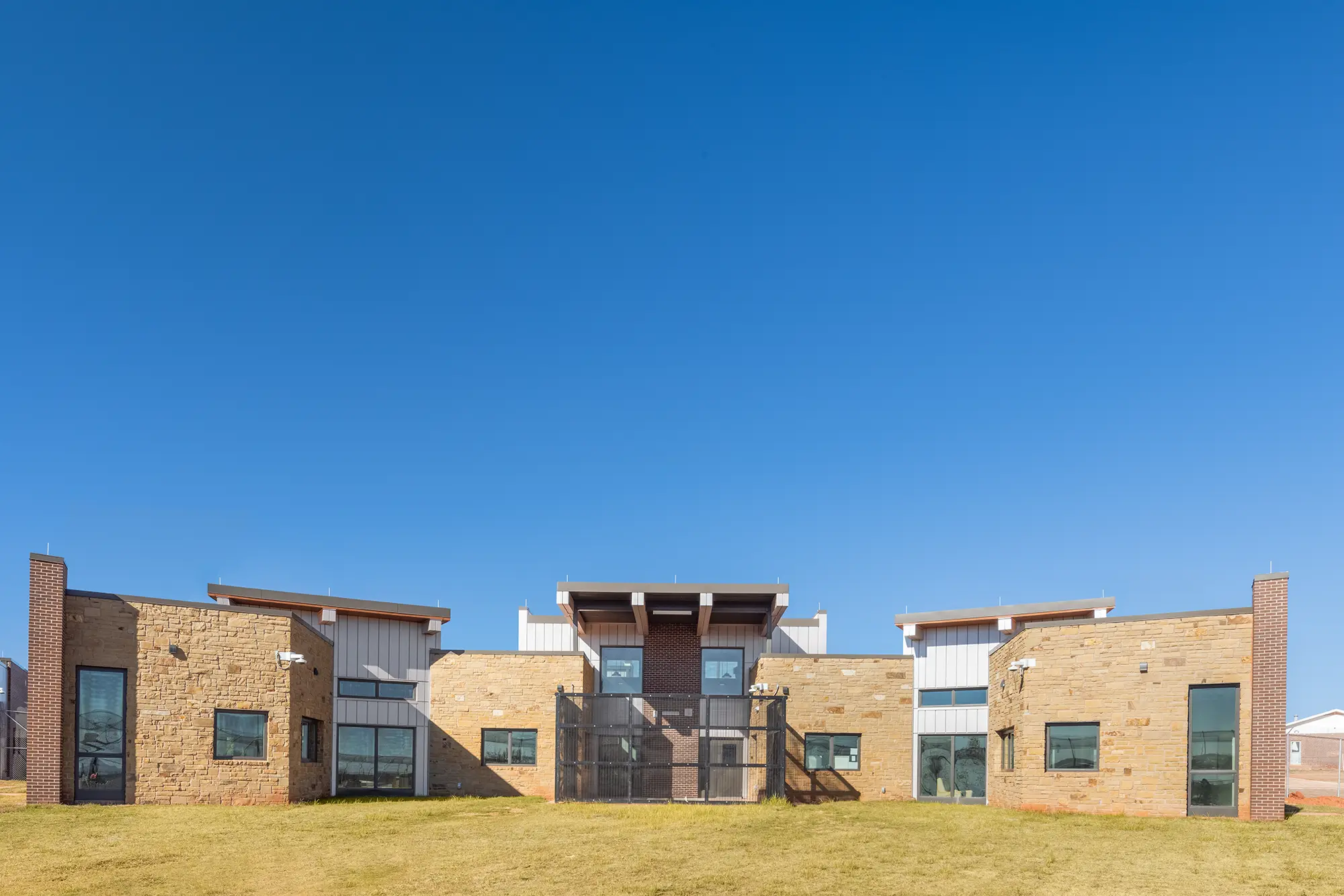 Modern building with brick and metal facade under clear blue sky. Features symmetrical design, large windows, and sits on a grassy lawn.