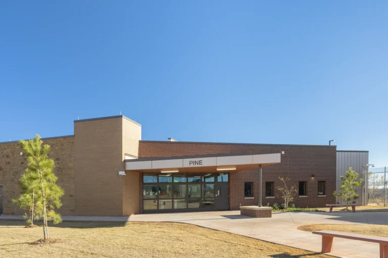 Modern brick building under a clear blue sky, labeled "Pine." The entrance is glass-paneled, surrounded by small trees and a concrete waModern brick building under a clear blue sky, labeled "Pine." The entrance is glass-paneled, surrounded by small trees and a concrete walkway. Calm, sunny setting.lkway. Calm, sunny setting.
