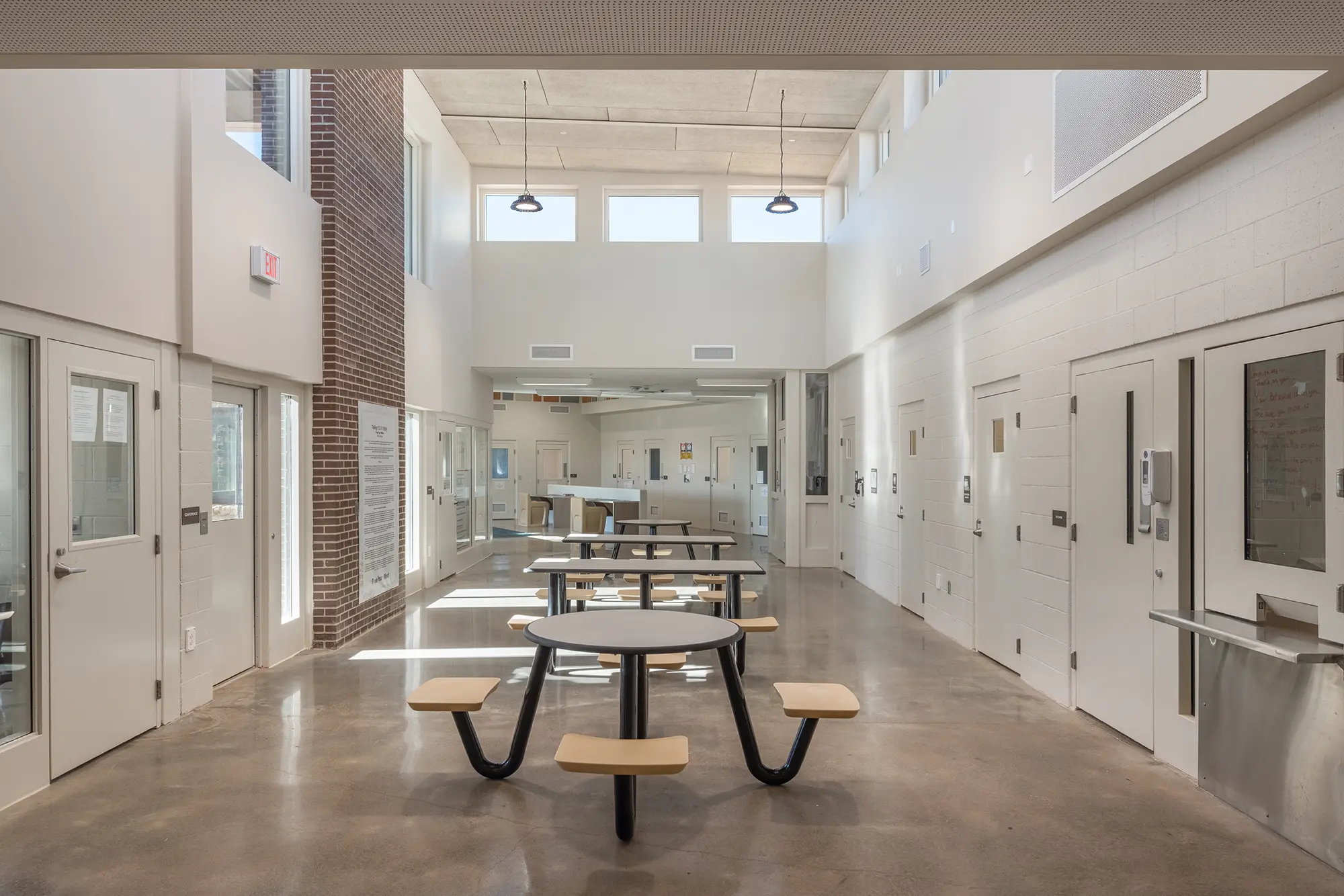 Spacious room with high ceilings, white walls, and multiple round tables with wooden benches. Natural light streams in through upper windows, creating a calm atmosphere.