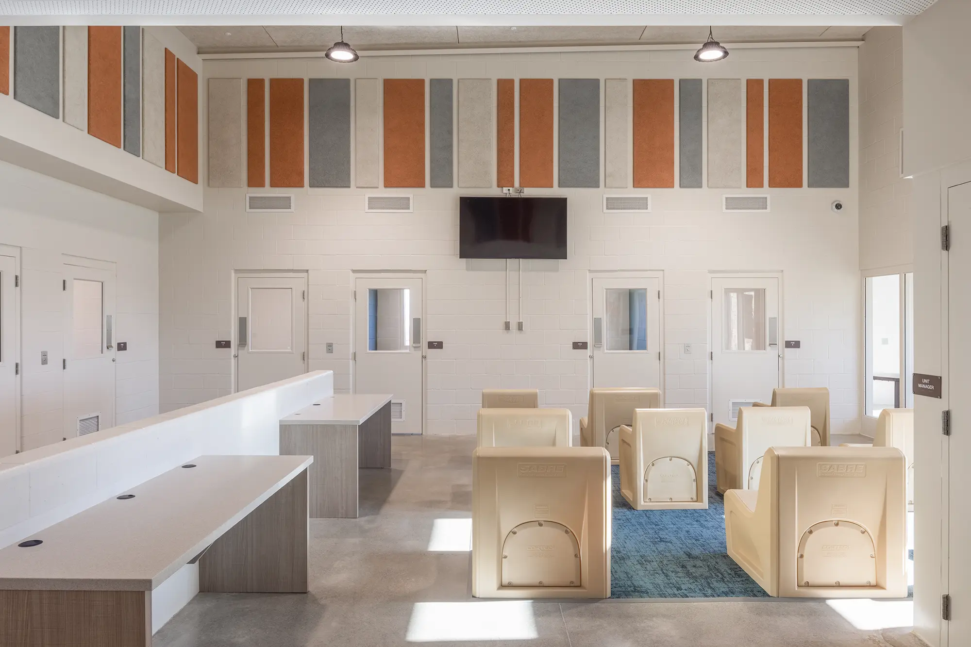 A bright, open waiting area features beige chairs, a blue rug, large TV on the wall, and colorful vertical panels. Sparse, modern, institutional feel.