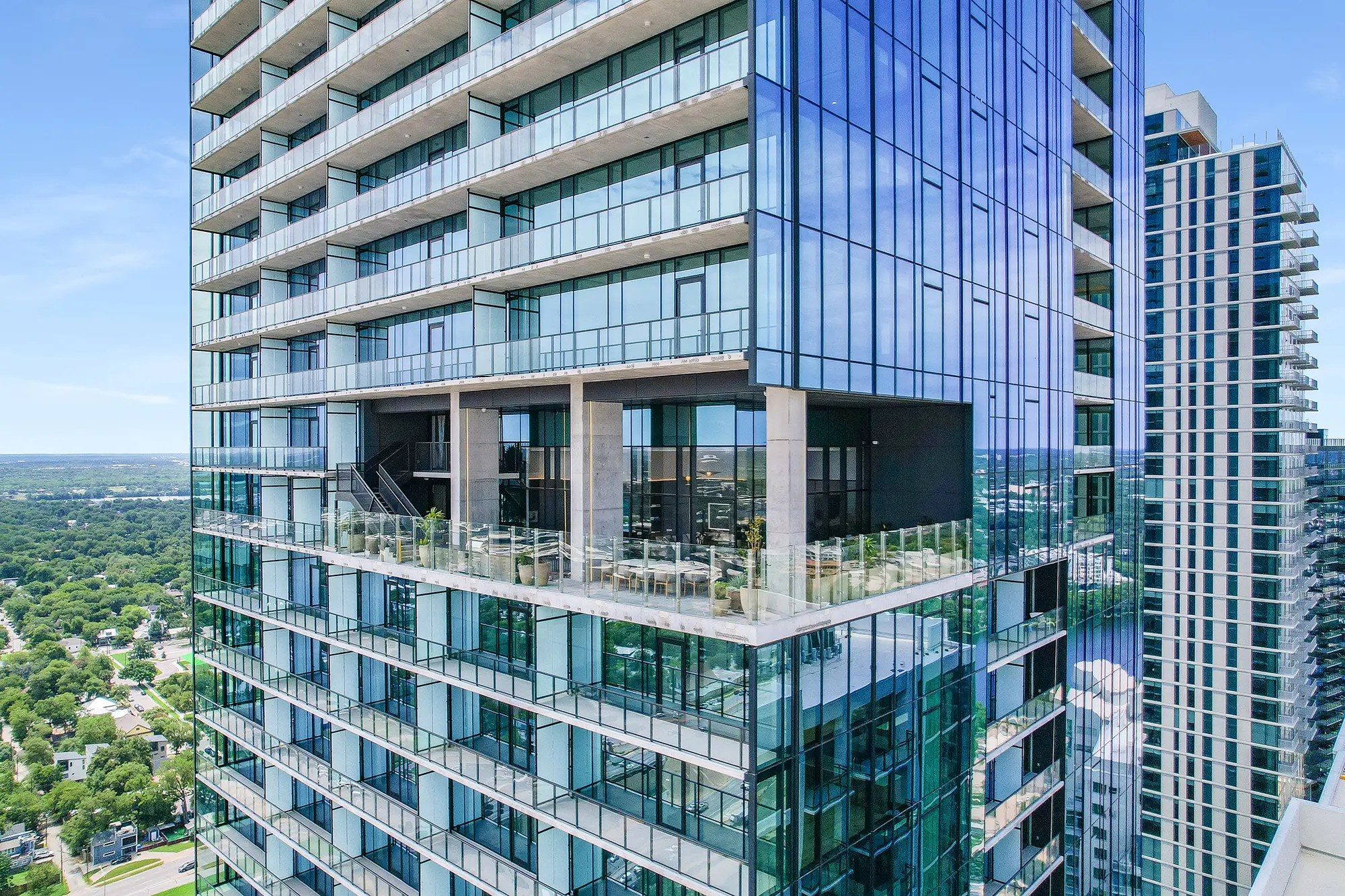 세련되고 현대적인 유리 고층 빌딩이 넓은 발코니와 야외 좌석을 갖추고 무성한 도시 풍경을 내려다보고 있다. 반사된 풍경은 고급스러움과 개방감을 전한다.