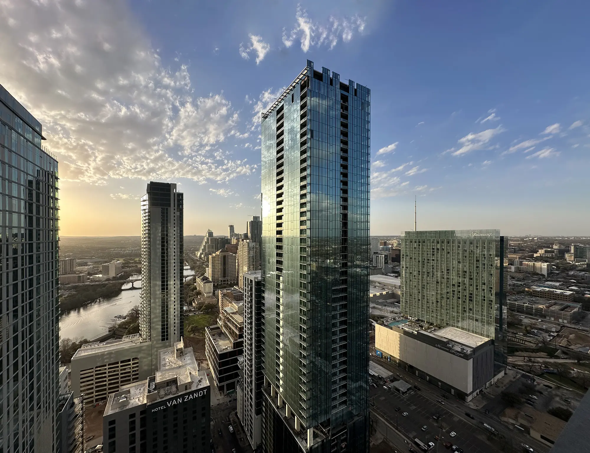 유리 외벽을 가진 고층 건물들이 석양 속 도시 풍경에 햇빛을 반사한다. 흐린 푸른 하늘 아래 배경으로 강이 흐르고 있다.