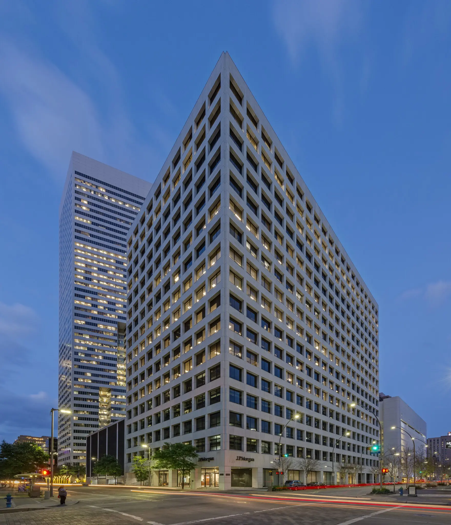 Tall office building at dusk, illuminated windows in a grid pattern. City street intersection below, faint car light trails, and a clear sky enhance the urban setting.