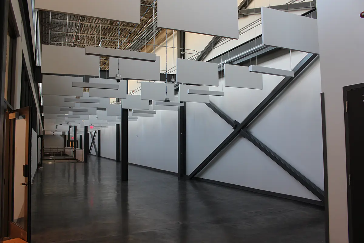 Spacious modern hallway with high ceilings, suspended white panels, and intersecting steel beams. The polished concrete floor adds an industrial feel.