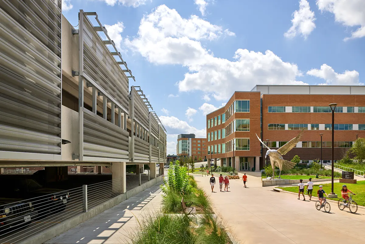 Modern campus scene with a brick building and a parking garage under a blue sky. Students walk and bike along a sunny pathway. Relaxed atmosphere.