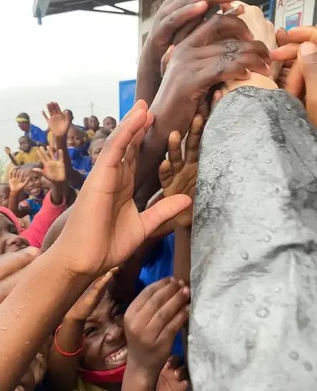 A group of joyful children and adults reach out with their hands, smiling and cheering under a rainy sky, creating a sense of community and excitement.