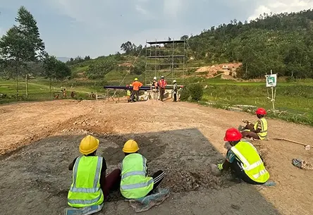Workers in yellow and red helmets and high-visibility vests sit on a cleared construction area in a rural landscape, overseeing a scaffolding setup. The atmosphere is industrious and collaborative.