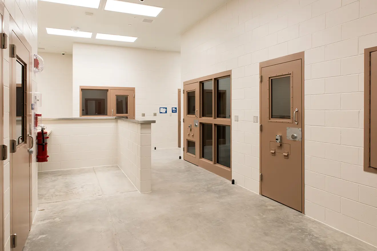 Interior of a pristine detention facility with white cinder block walls, tan steel doors with small windows, a glass-enclosed office, and a fire extinguisher.