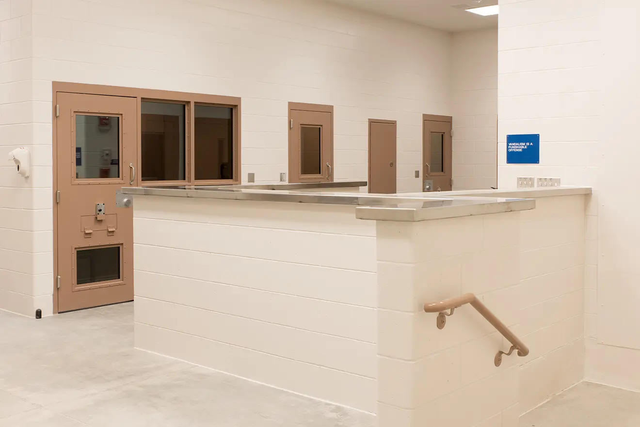 A sterile room with white brick walls features brown metal doors with small windows, a countertop, and a handrail. The atmosphere is utilitarian and stark.