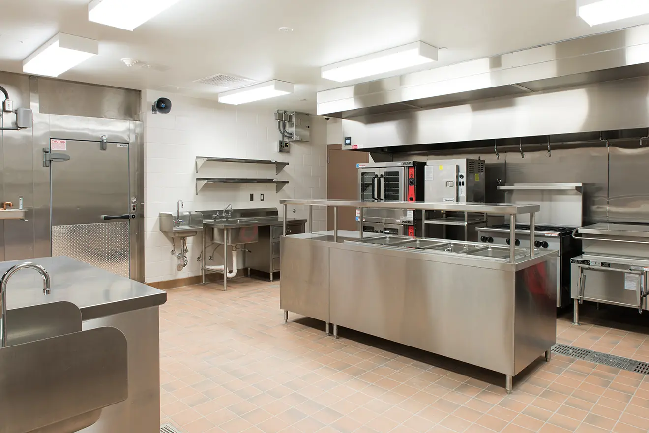 Commercial kitchen with stainless steel appliances, including ovens, sinks, and shelves. The space is clean and organized, evoking a professional tone.