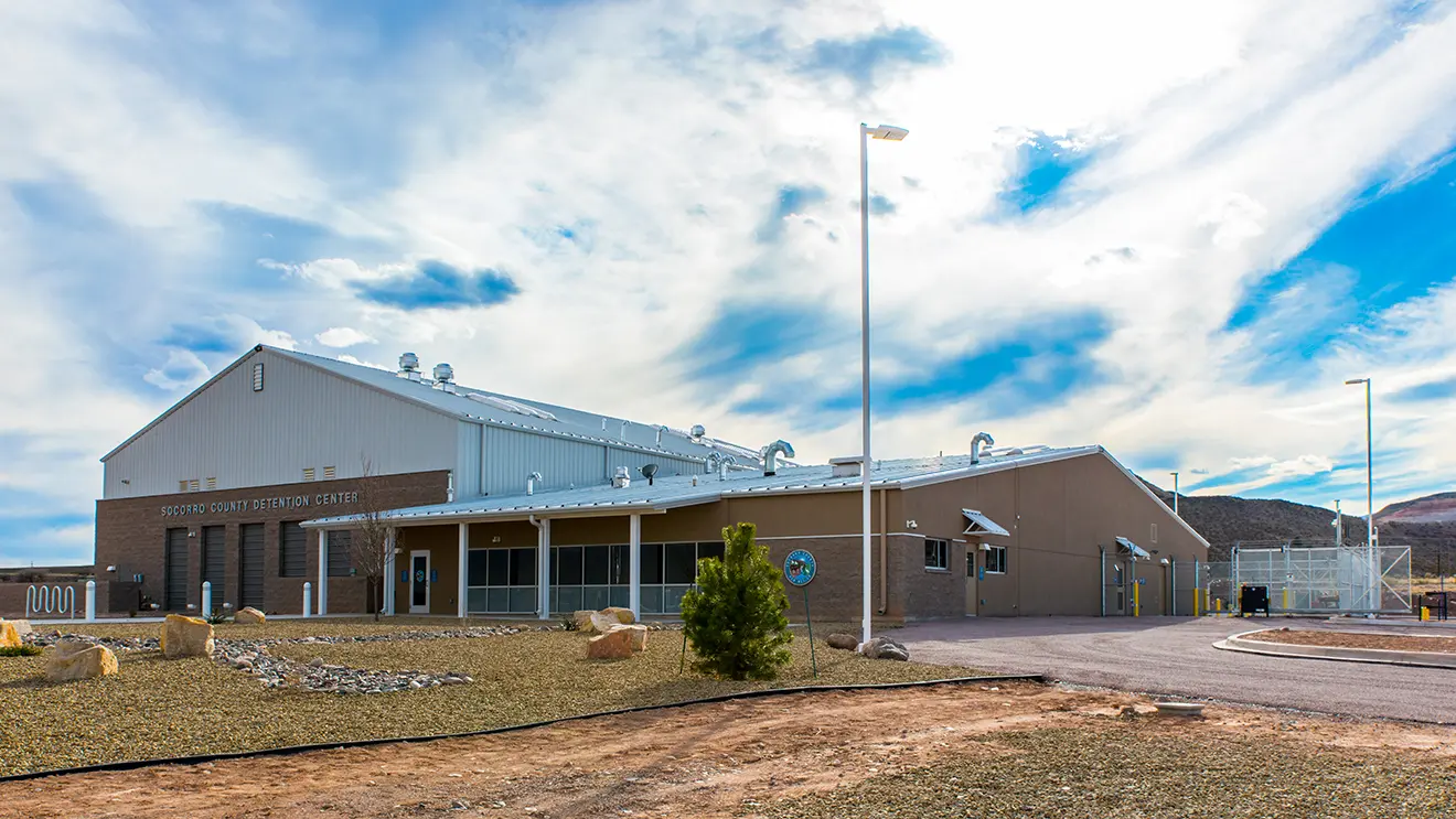 A large, modern detention center stands against a bright sky with scattered clouds. The building is surrounded by a rocky landscape, evoking a sense of isolation.