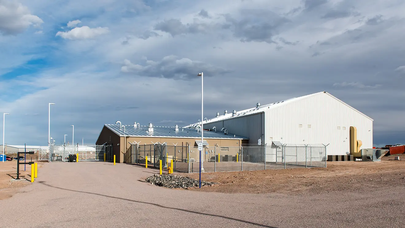 A large industrial building with a fenced perimeter sits under a cloudy sky. The surroundings are arid with dirt roads and sparse vegetation.