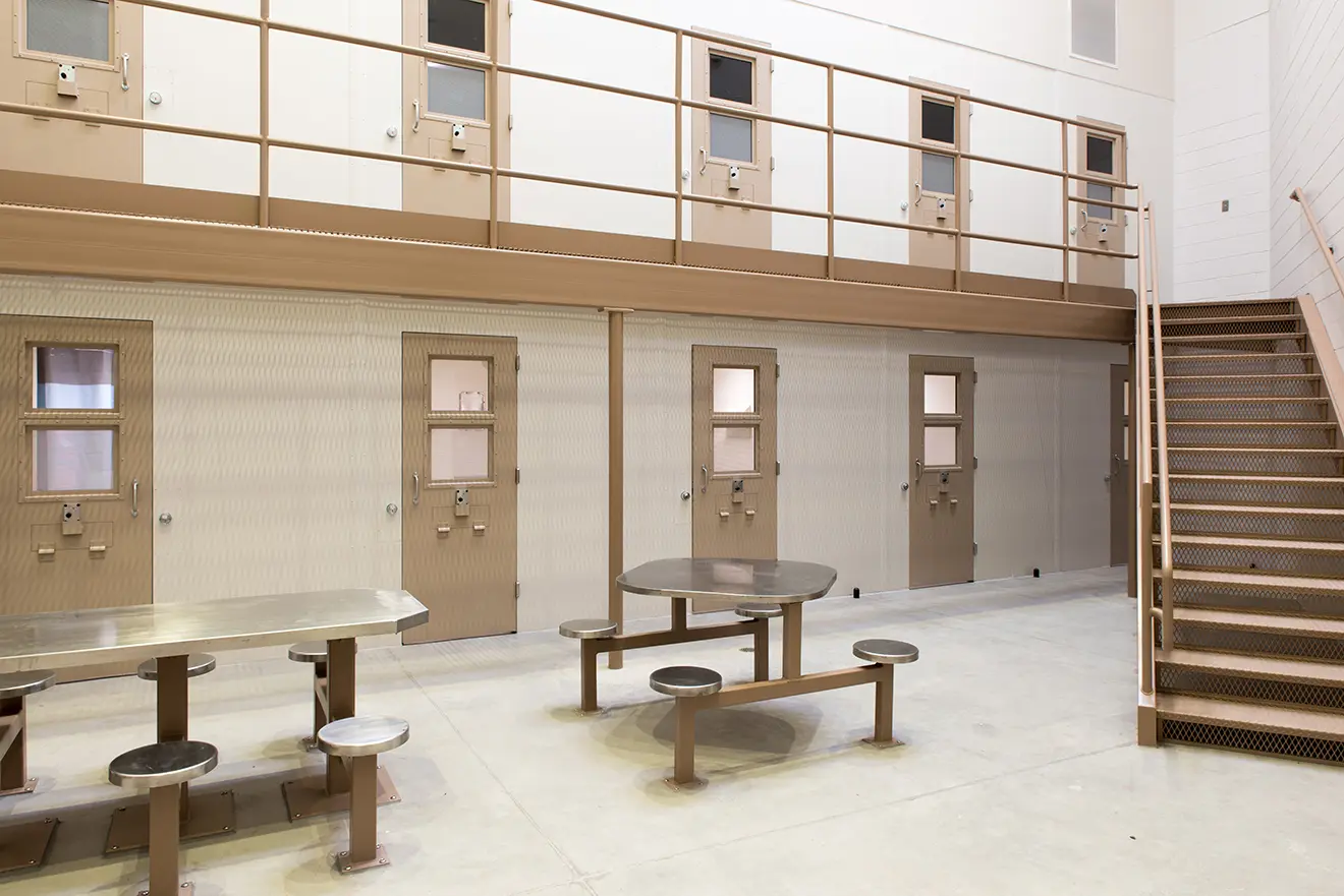 Interior of a prison block with beige cell doors and a second-floor balcony. Metal tables and chairs are on a concrete floor, creating a stark, utilitarian atmosphere.